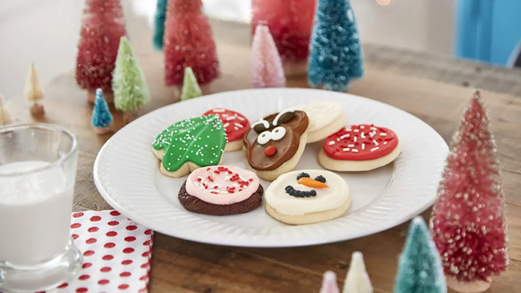 History of Christmas cookies with a plate of decorated cookies on a table surrounded by small fake trees.