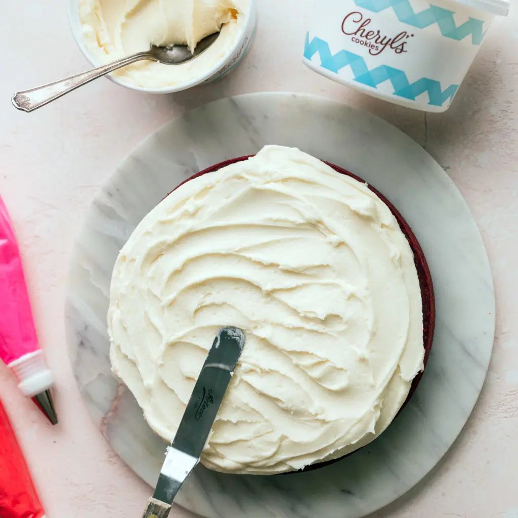 Red velvet cake recipe with a half iced cake with tubs of Cheryl's buttercream frosting next to it.
