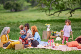 A photo of picnic with a group of people sitting outside on a blanket surrounded by food and gifts