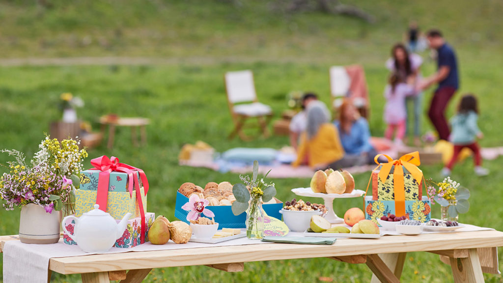 A photo of picnic with an outdoor table full of gifts and food with a group of people behind it sitting on a blanket.